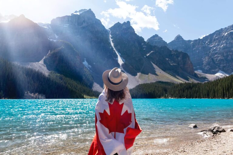 a man wearing a hat and sitting on a beach with a body of water and mountains in the